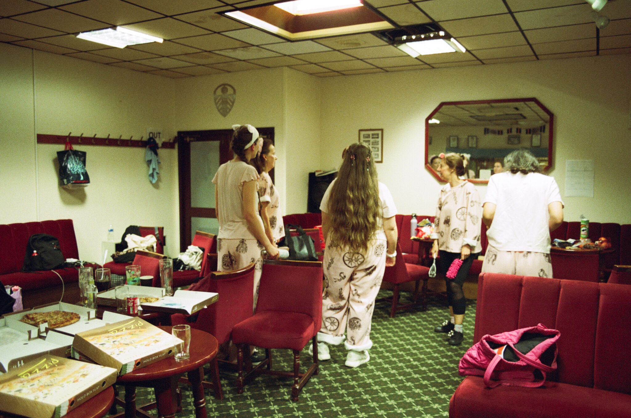 A group of performers wearing coordinated pale pink costumes gather in a backstage dressing room before or after a performance. Pizza boxes, drinks and personal belongings are scattered around the room, creating a relaxed, informal atmosphere as the group talks and prepares together.