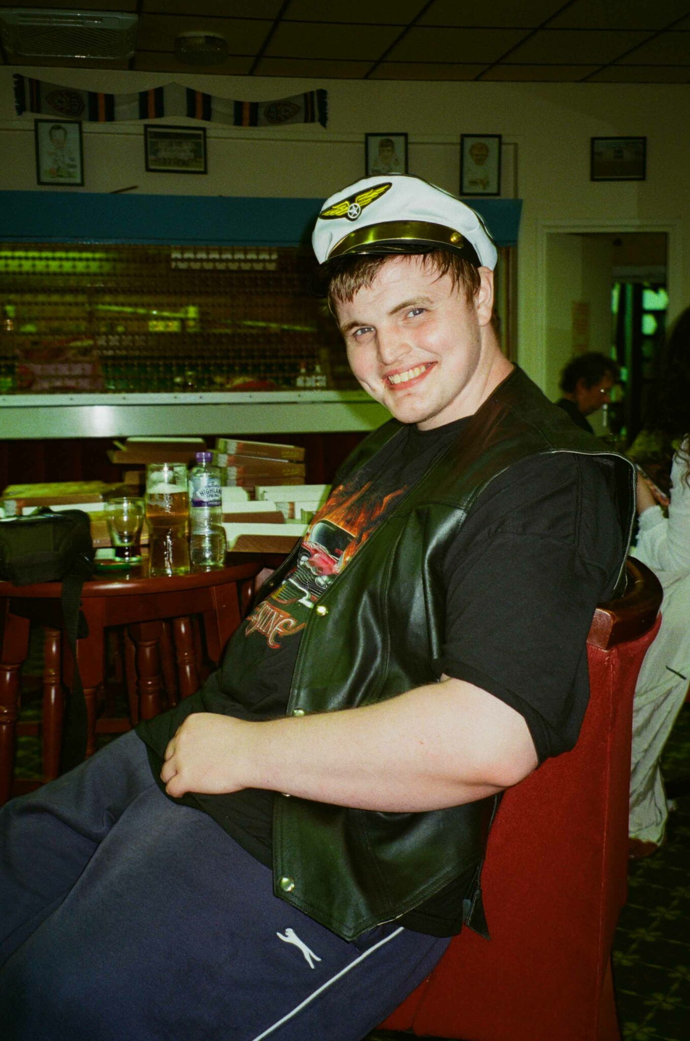 A person wearing a white captain's hat, black leather waistcoat and graphic T-shirt sits smiling at a table in a community venue. Drinks, pizza boxes and the bar in the background create a relaxed atmosphere between performances at the cabaret event.