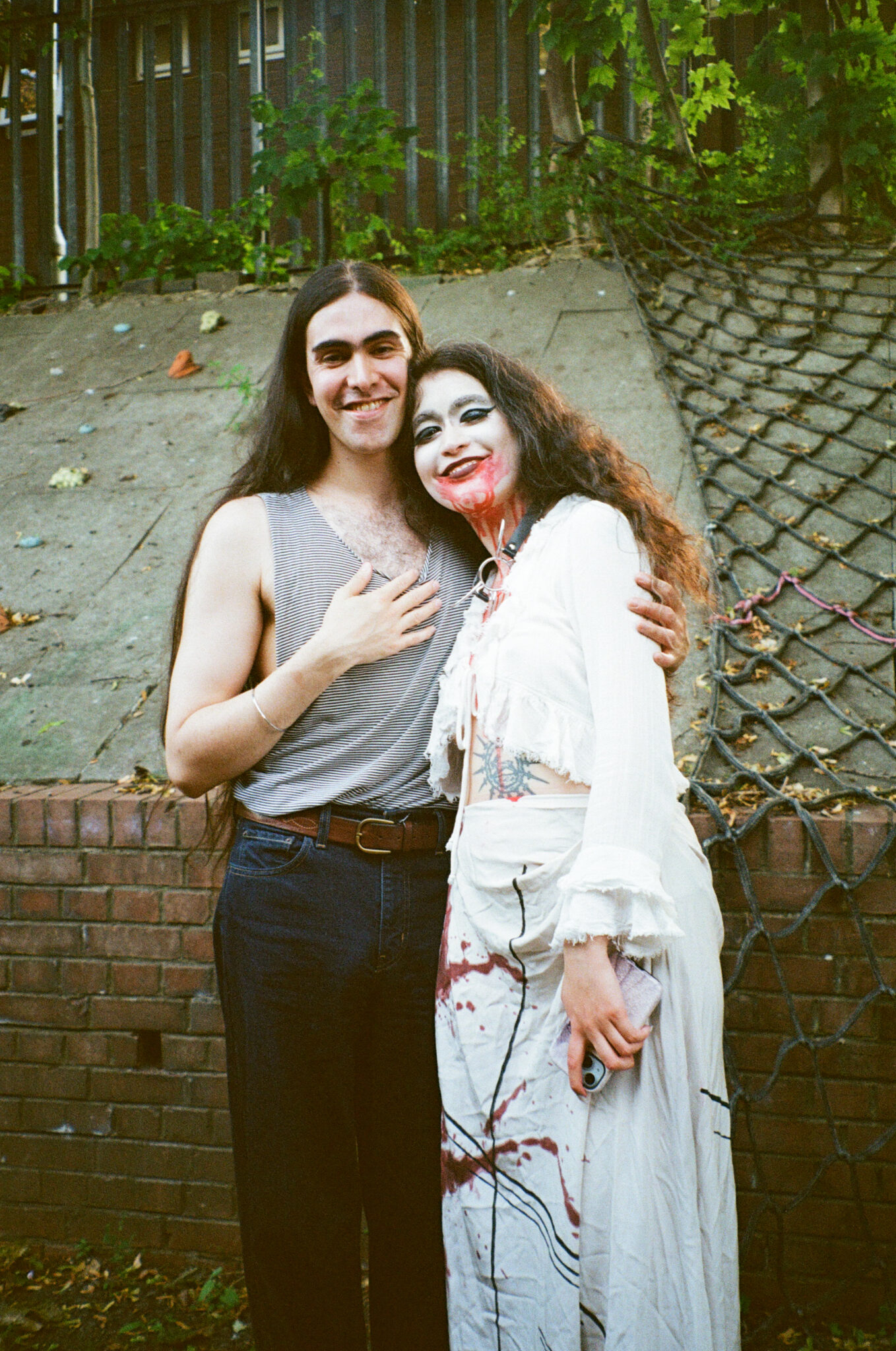 Two people stand arm in arm outdoors, smiling towards the camera after a performance. One wears a flowing white costume with dramatic theatrical makeup featuring white face paint and vivid red details around the mouth and neck, while the other is dressed casually. The portrait captures a warm moment of friendship and celebration following the community cabaret event.