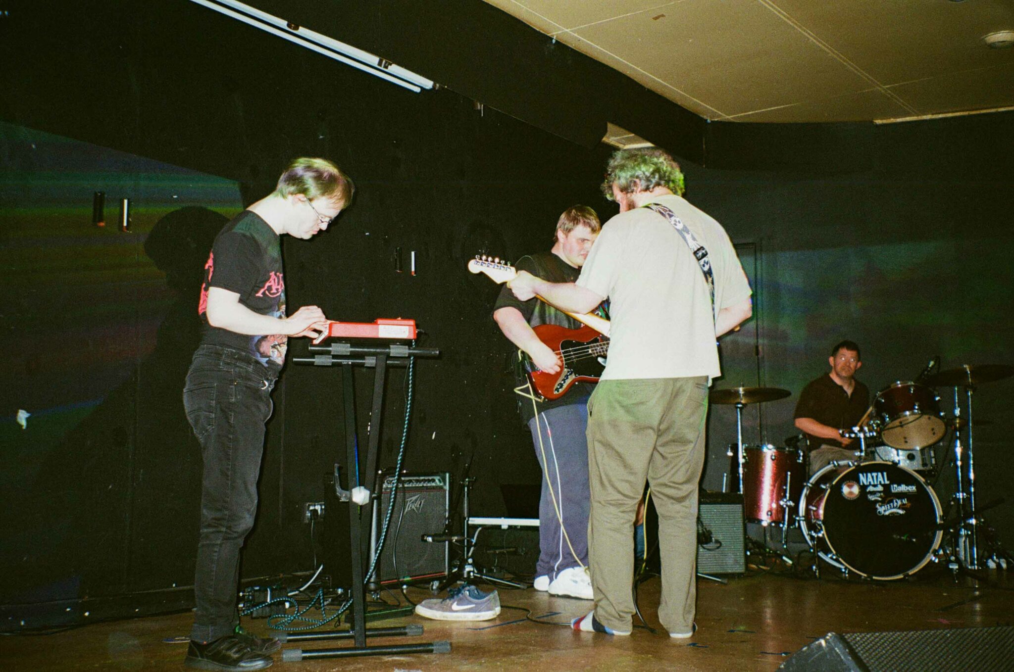 4 people perform on stage with guitars, drums and keyboard. The stage bright with a black curtain behind and the feel of the music performance is rock.