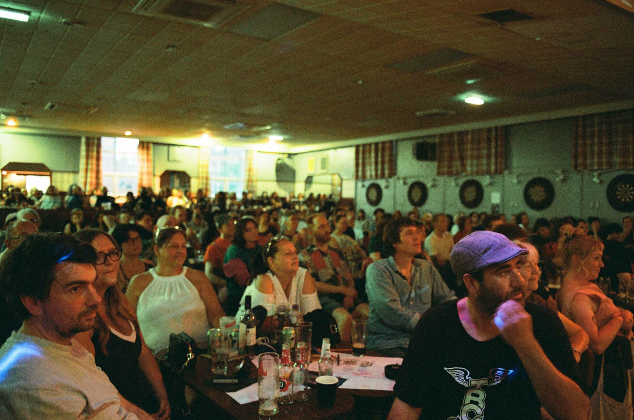 Audience seated at tables in a community hall watching a live performance during Cabaret Mystic, with drinks on tables and warm stage lighting illuminating the room.