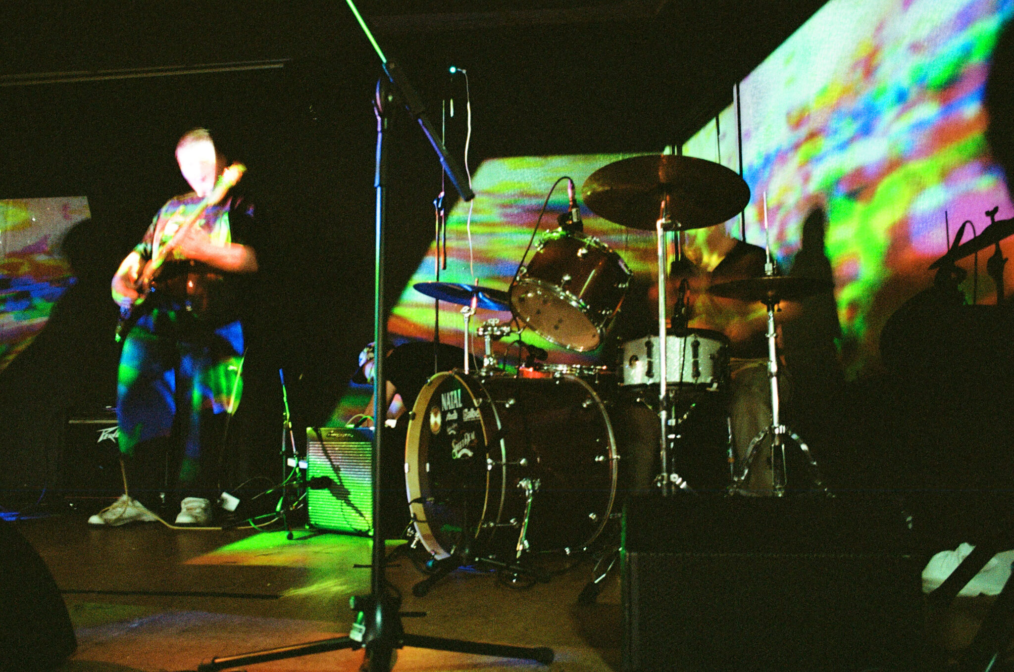 people perform on stage with guitars, drums and keyboard. The stage is mostly in shadow but it is illuminated by disco line giving a bright colored streak of light going across the stage.