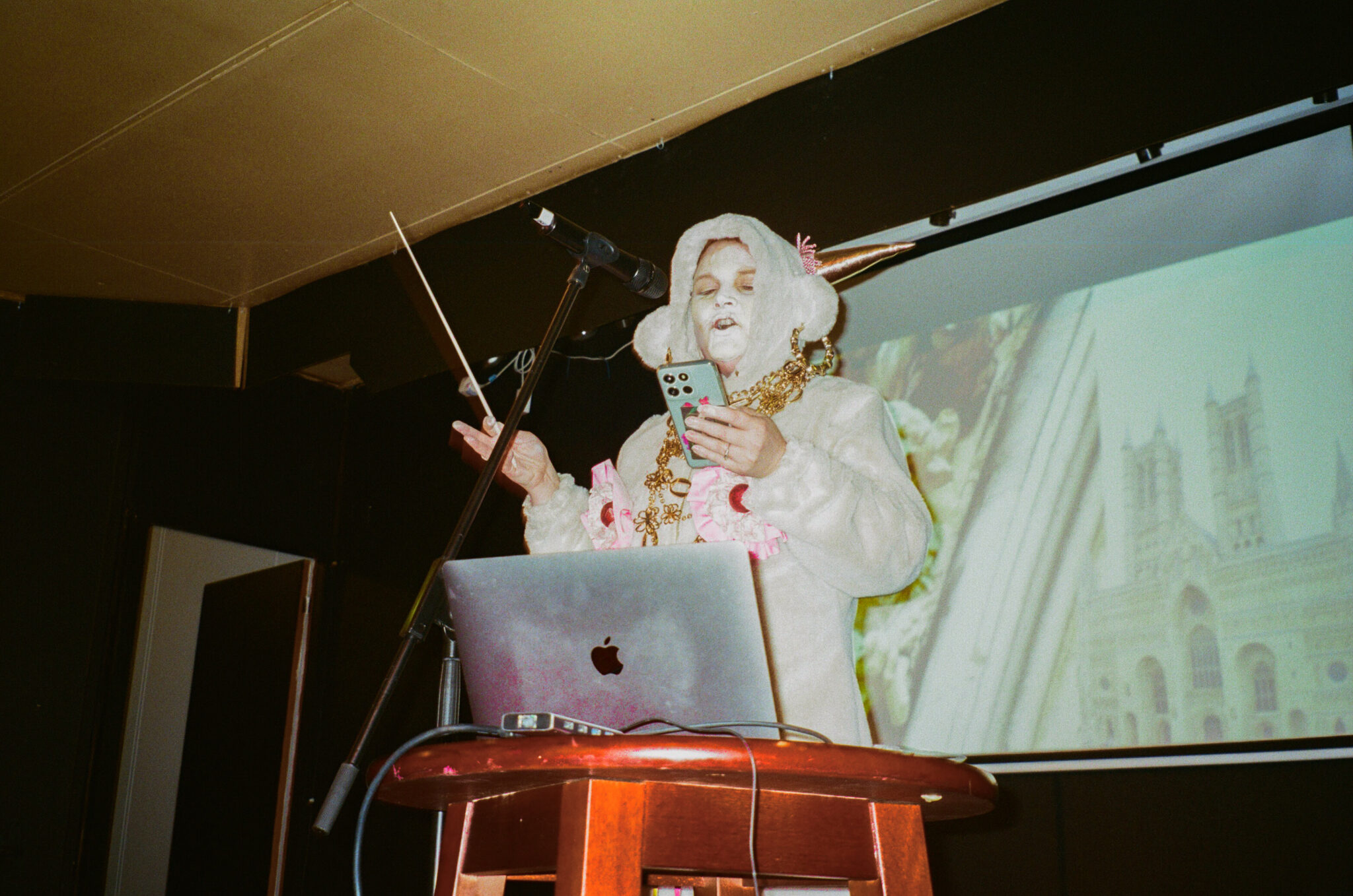 A performer in an elaborate white costume with oversized ear-like headpieces, gold jewellery and theatrical makeup stands at a microphone, reading from a smartphone while holding a sheet of paper. A laptop sits on the table in front of them, with a projected image on the screen behind, suggesting a multimedia spoken-word or performance art event.