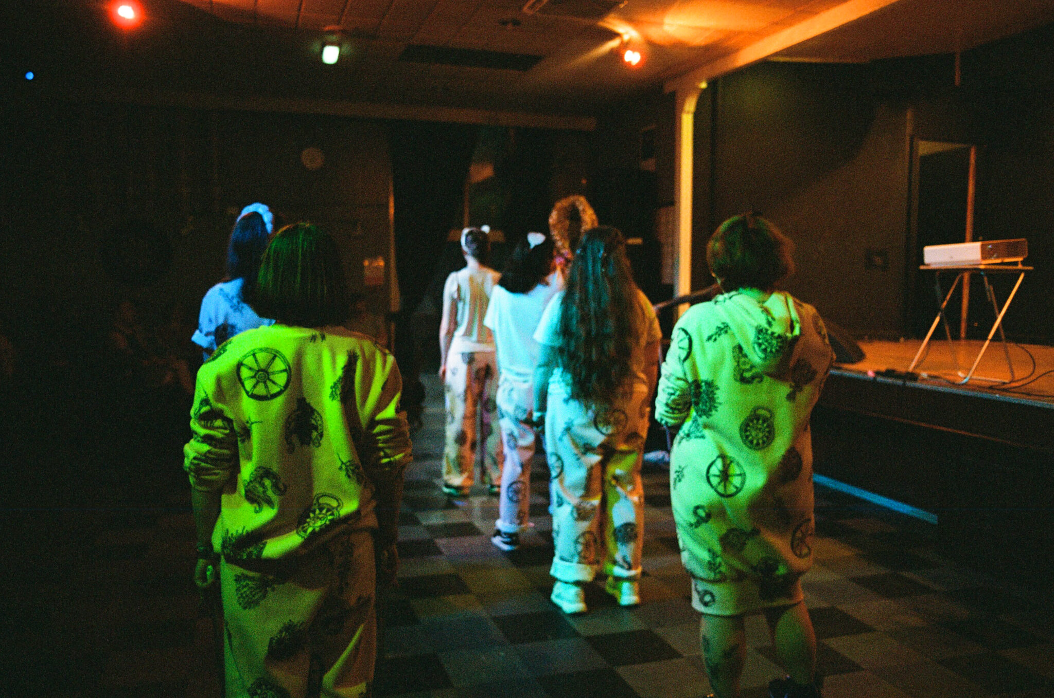 A group of performers wearing coordinated pale pink costumes walk in single file across the floor of a dimly lit community hall. Coloured stage lights cast green and orange tones over the scene as they move away from the camera towards the performance space, suggesting a choreographed procession as part of the live event.