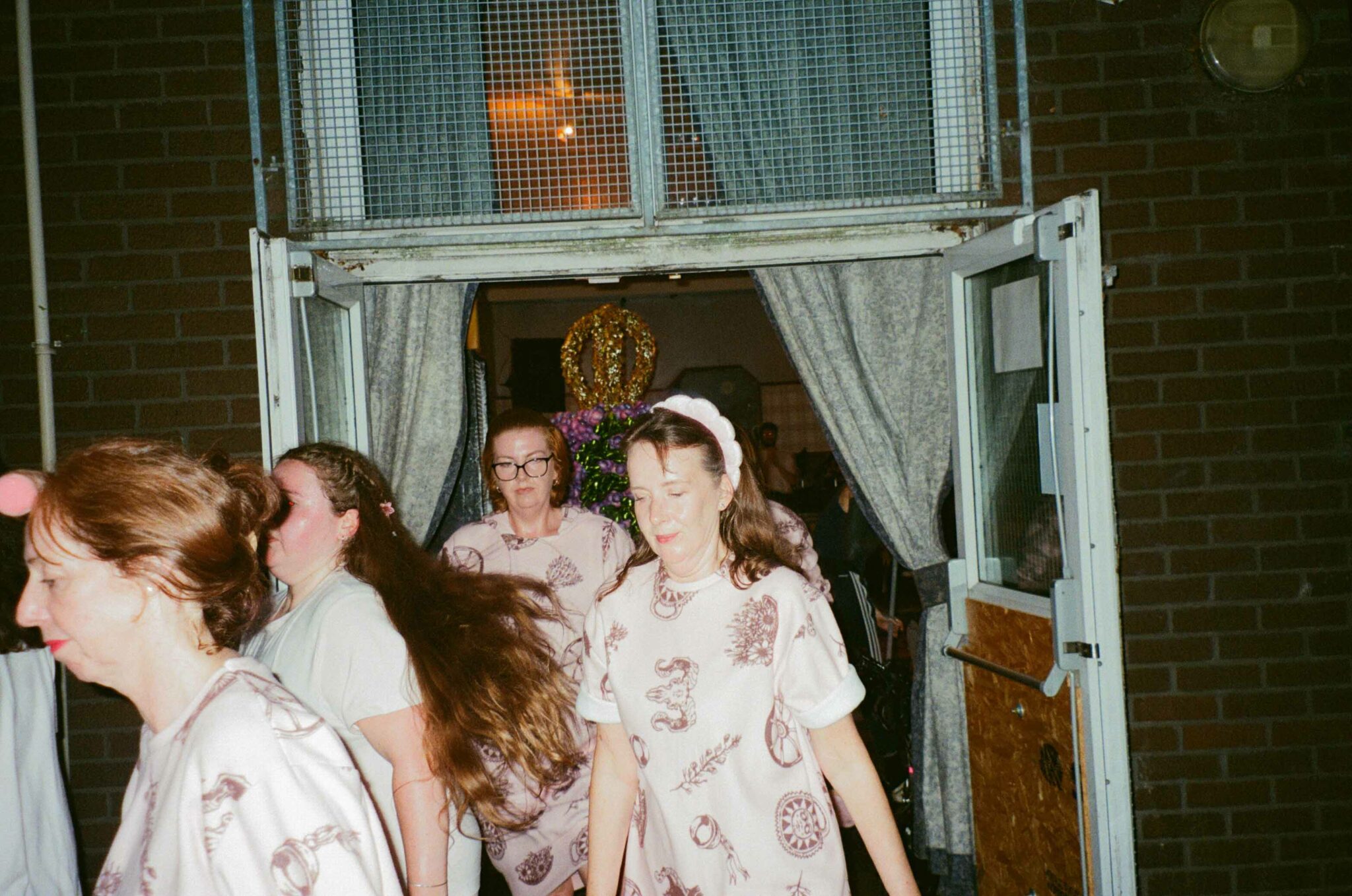 A group of performers wearing coordinated pale outfits with printed designs walk through an open doorway into an outdoor space. Curtains frame the entrance, while a decorative floral sculpture topped with a gold crown is visible inside the building, suggesting a procession or transition as part of a live performance.