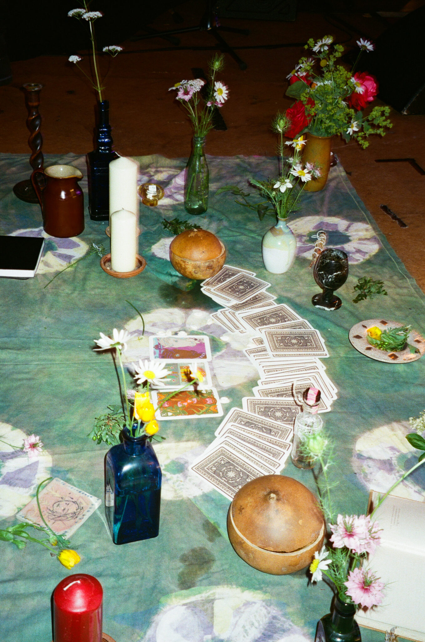 A ceremonial arrangement on the floor combines wildflowers in glass bottles, candles, bowls, playing cards, books and other small objects on a patterned cloth. The carefully arranged display suggests a ritual or participatory installation prepared as part of the live arts event.