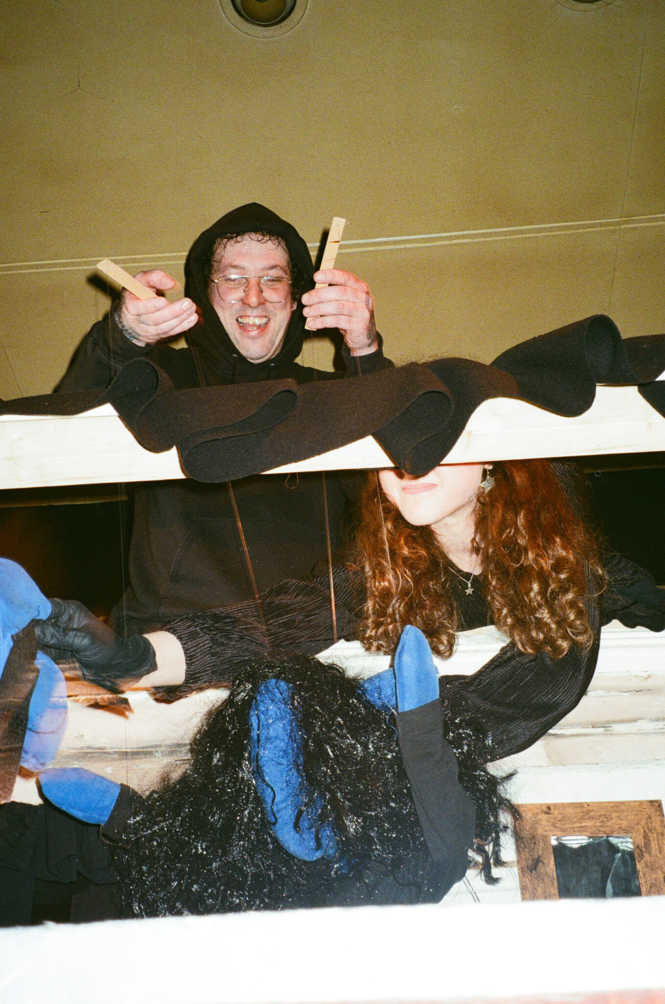 Two puppeteers dressed in black operate a puppet theatre set around a white wooden fireplace, manipulating a black puppet with bright blue hands and feet using strings and wooden controls. The close-up view reveals the craftsmanship of the live puppet performance and the performers working together behind the scenes.