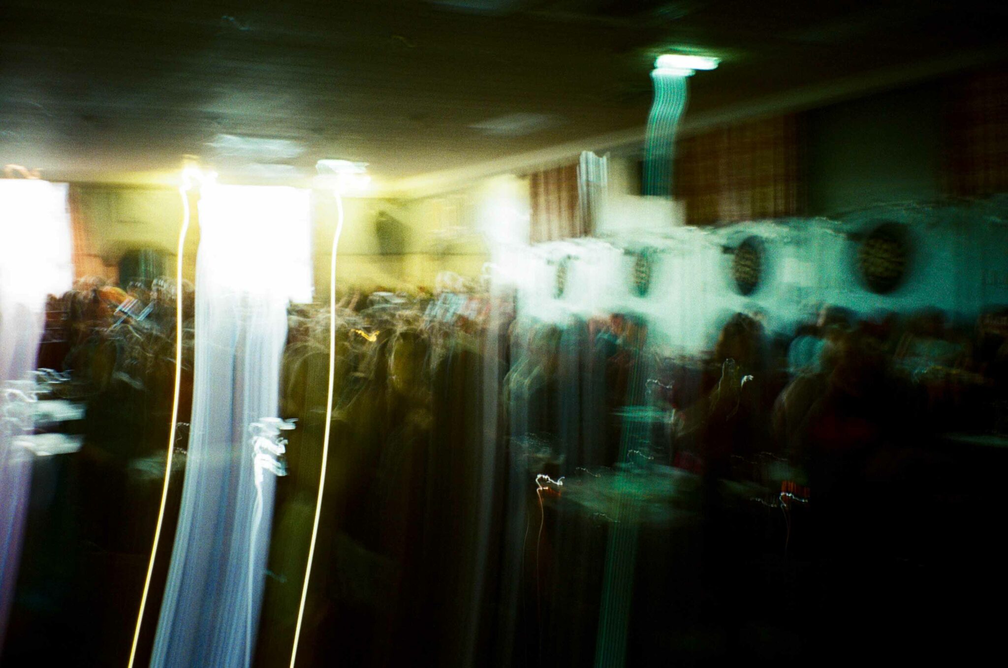 An intentionally blurred photograph of a crowded indoor gathering, with long streaks of light created by camera movement. Bright white and green lights glow across the dimly lit room, while the figures of people, tables, and dartboards are rendered as soft, abstract shapes, conveying the energy and atmosphere of the event rather than individual details.