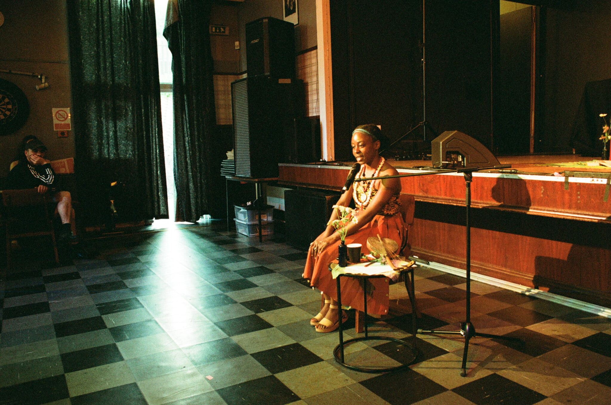 A person sits beside a small table, speaking into a microphone in a community hall with a low stage behind them. Warm light illuminates the speaker and a vase of flowers on the table, while audience members sit around the room, creating an intimate atmosphere for a spoken-word performance or conversation.