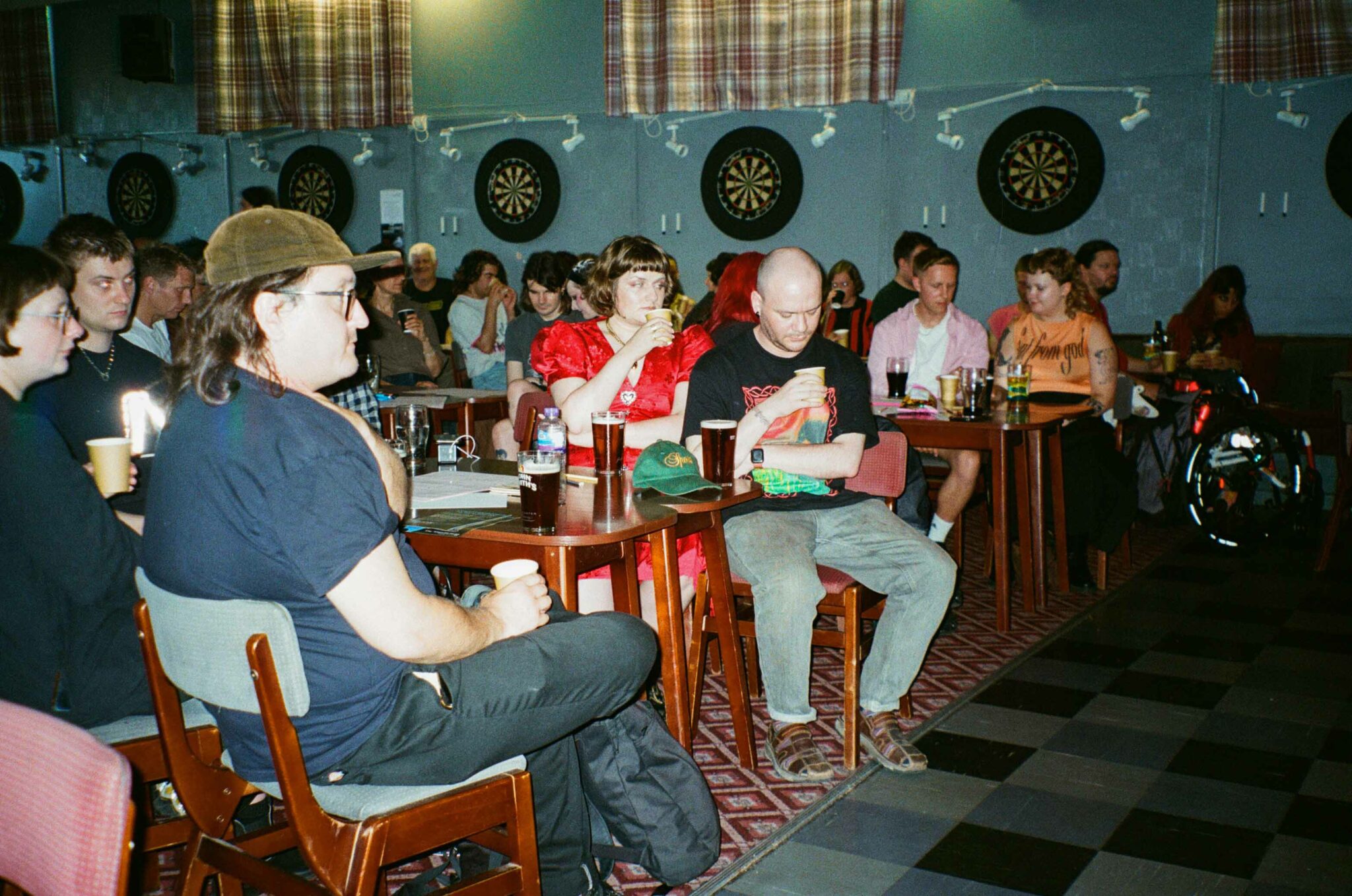 Audience members sit at tables in a community hall, watching a live performance while holding drinks in paper cups and glasses. The informal cabaret setting, with dartboards on the walls and people seated throughout the room, creates a relaxed, sociable atmosphere.