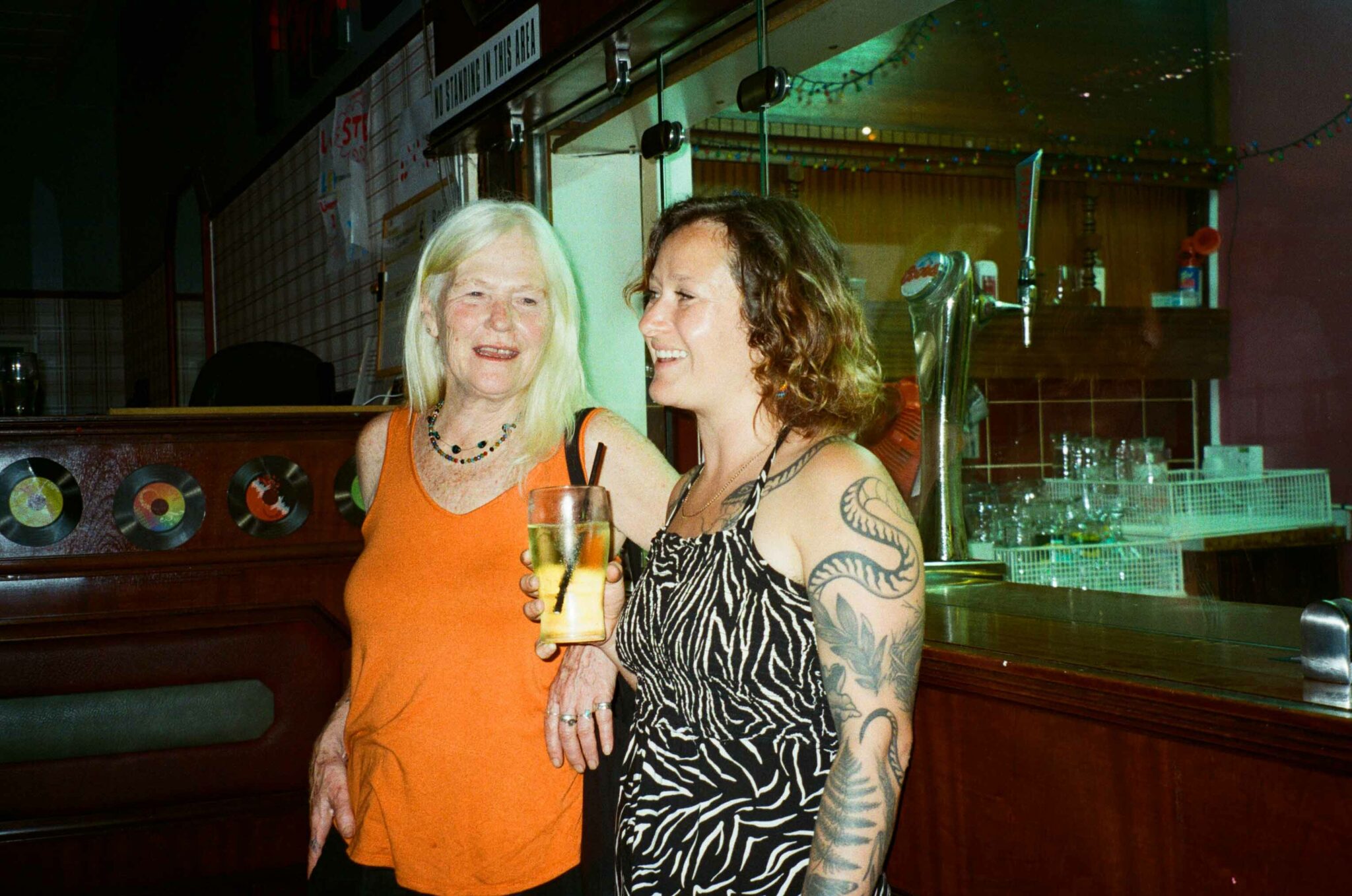Two people stand smiling and talking beside the bar in a community venue. One holds a drink while they chat, capturing a friendly moment between performances at the event.