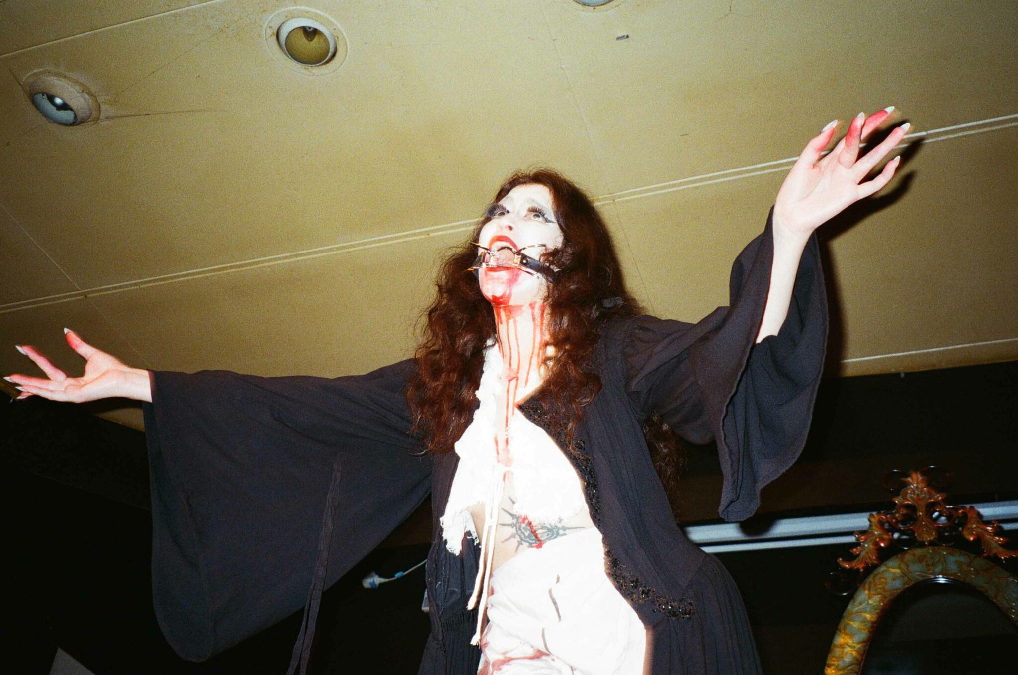 A person with long black hair in dramatic white face paint makeup with a ghostly appearance stands with arms raised, evoking a haunting and eerie atmosphere. They wear a dark robe and have theatrical fake blood on their face.