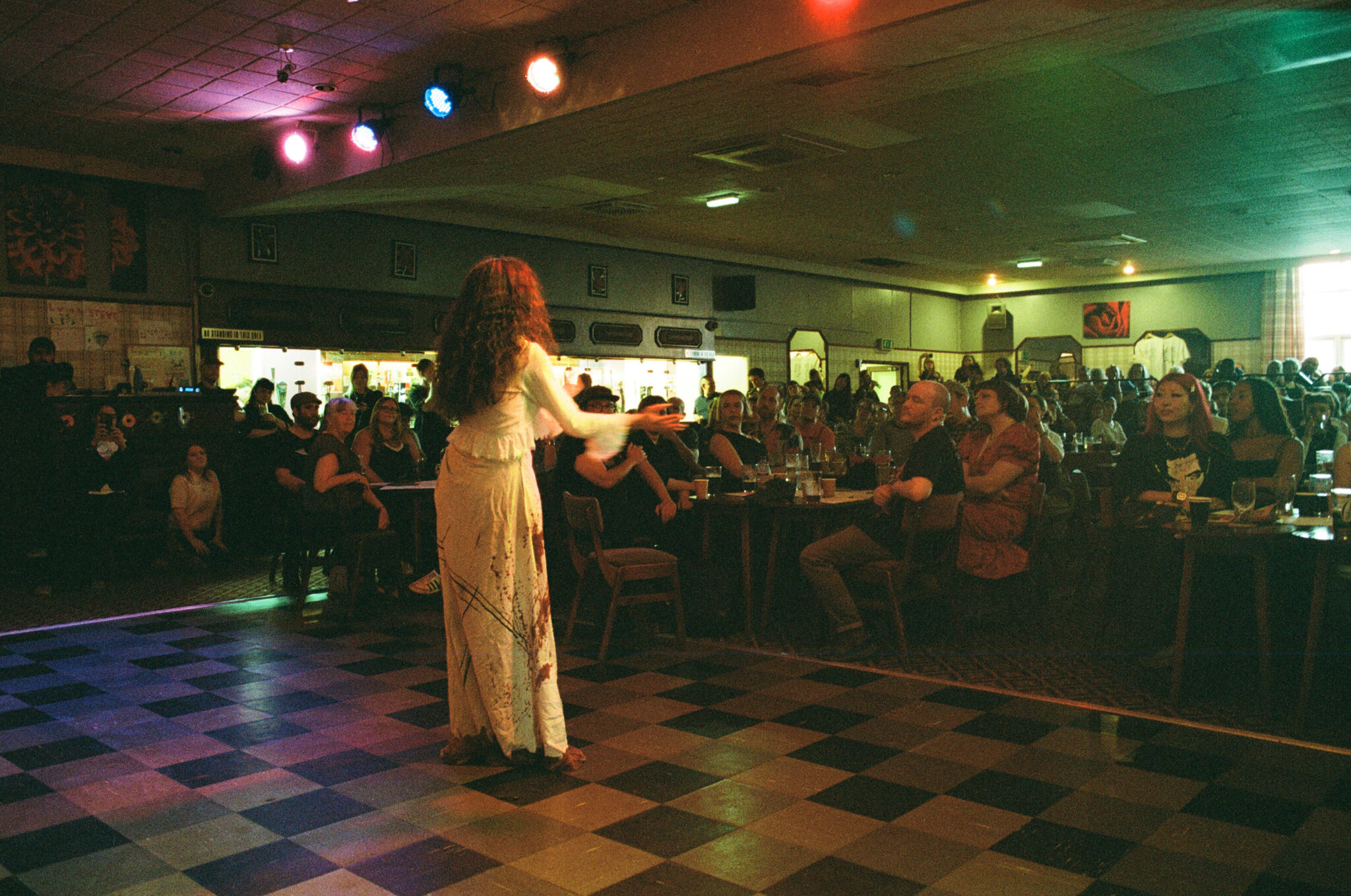 A person with long black hair in a long dress stands on a checkered stage facing an audience seated at tables in a dimly lit venue with colored spotlights overhead.