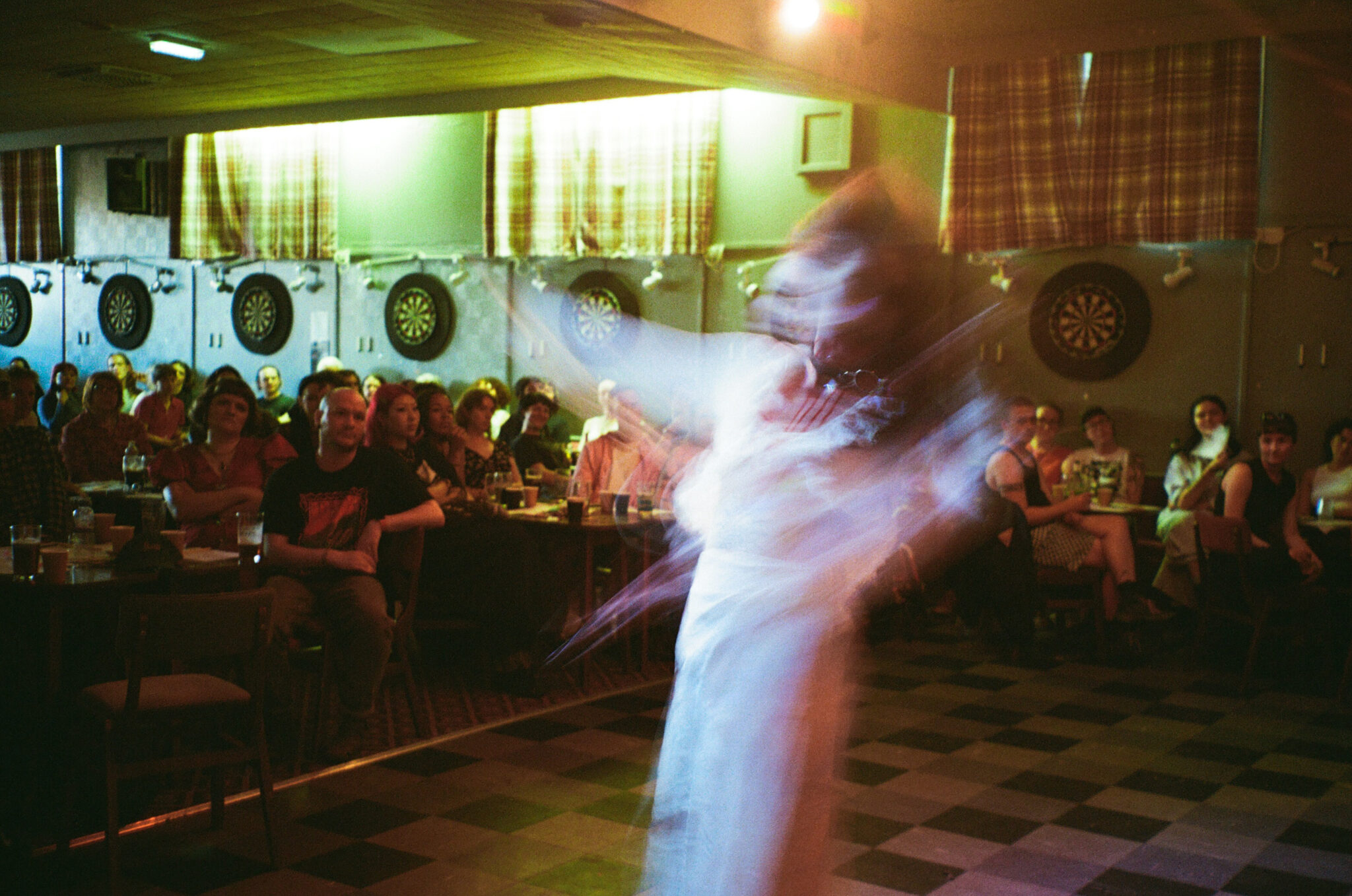An intentionally blurred photograph of a performer moving across the floor of a community hall while an audience watches from tables around the room. Motion blur creates flowing white shapes that convey the energy of the performance, with dartboards and warm lighting visible in the background.