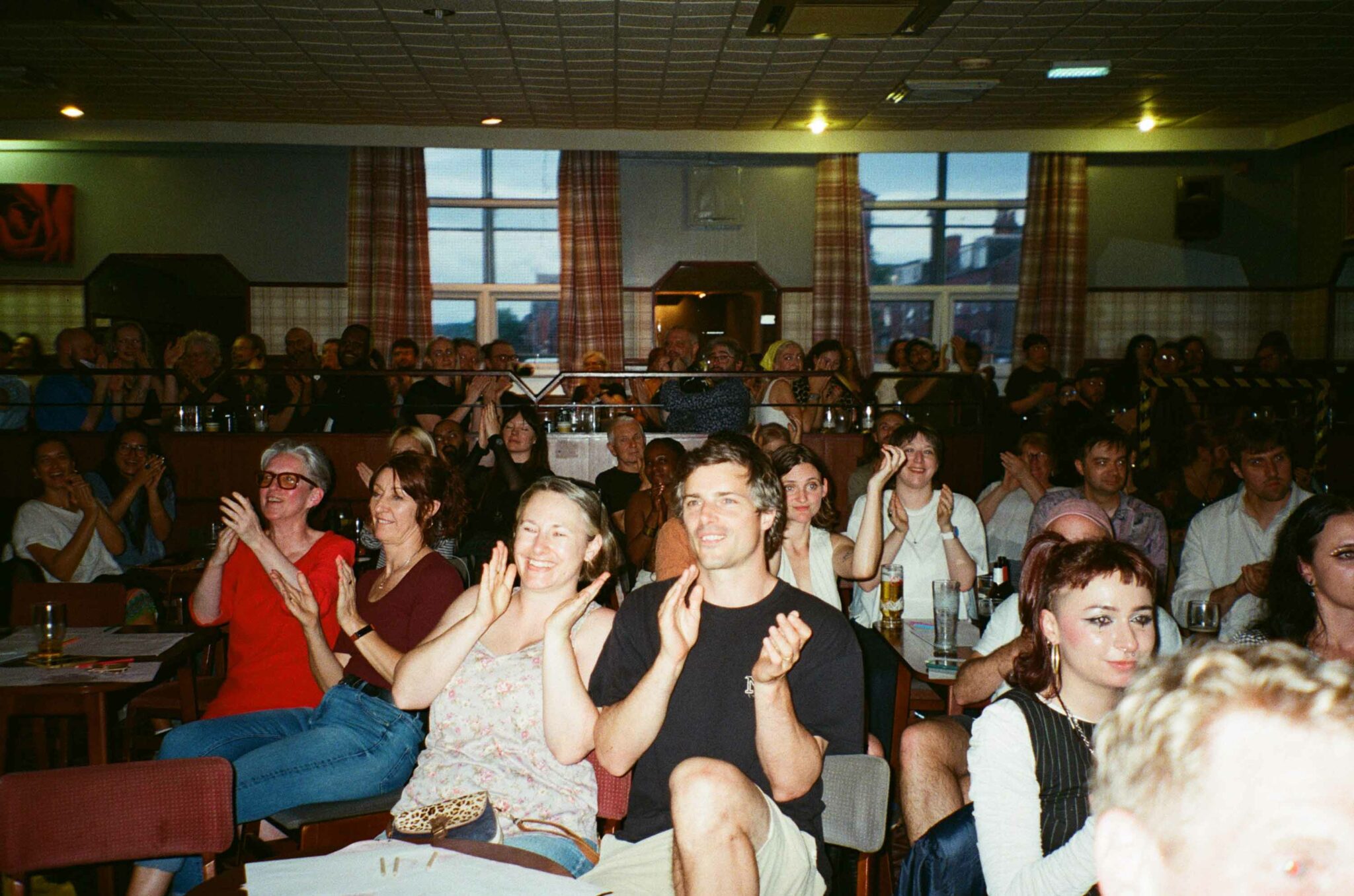 Audience members fill a community hall, smiling, clapping and applauding at the end of a live performance. Seated at tables with drinks, the crowd creates a warm, celebratory atmosphere that reflects the shared enjoyment of the community cabaret event.