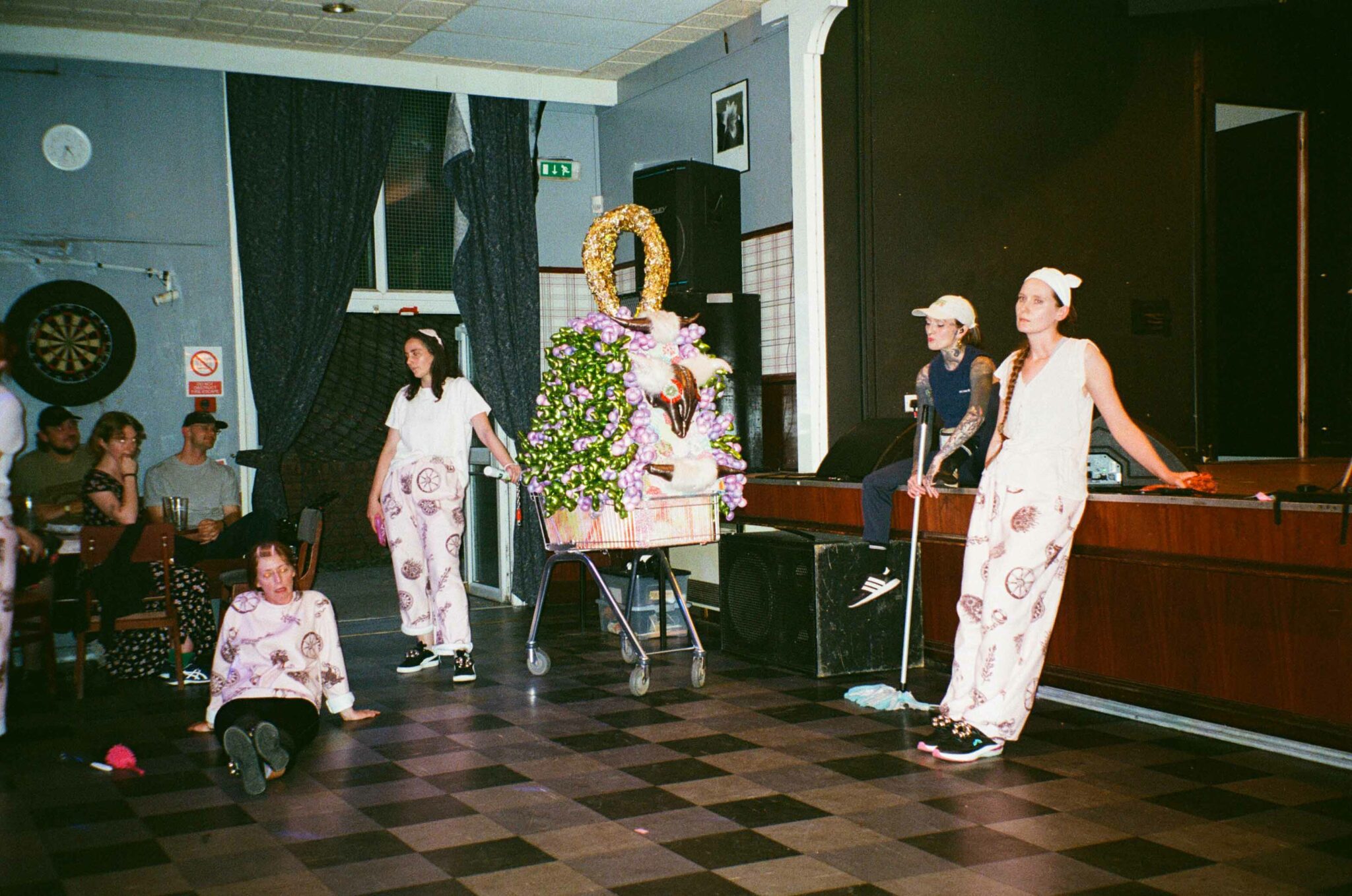 A group of performers occupy the floor of a community hall during a live performance. One sits on the floor while others stand around a decorated trolley carrying a colourful sculptural arrangement, as audience members watch from the side. The performers wear coordinated white outfits with printed designs, creating a playful, theatrical scene.