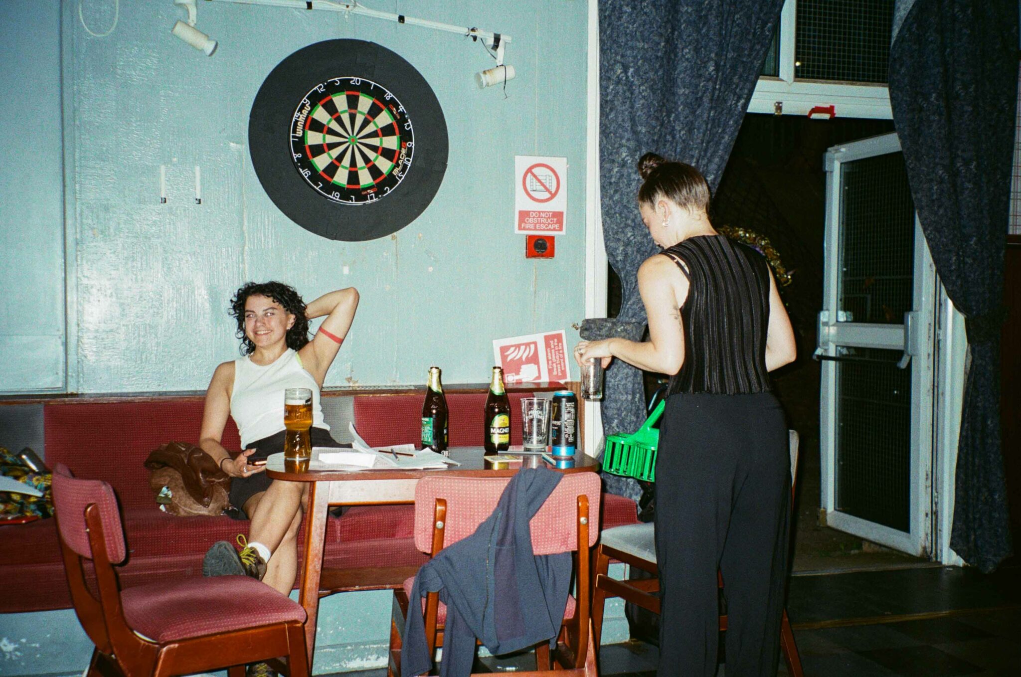 Two people chat beside a table in a community hall during an arts event. One relaxes on a bench with a drink while the other stands nearby holding a glass, with dartboards, an open doorway and empty bottles adding to the informal social atmosphere.