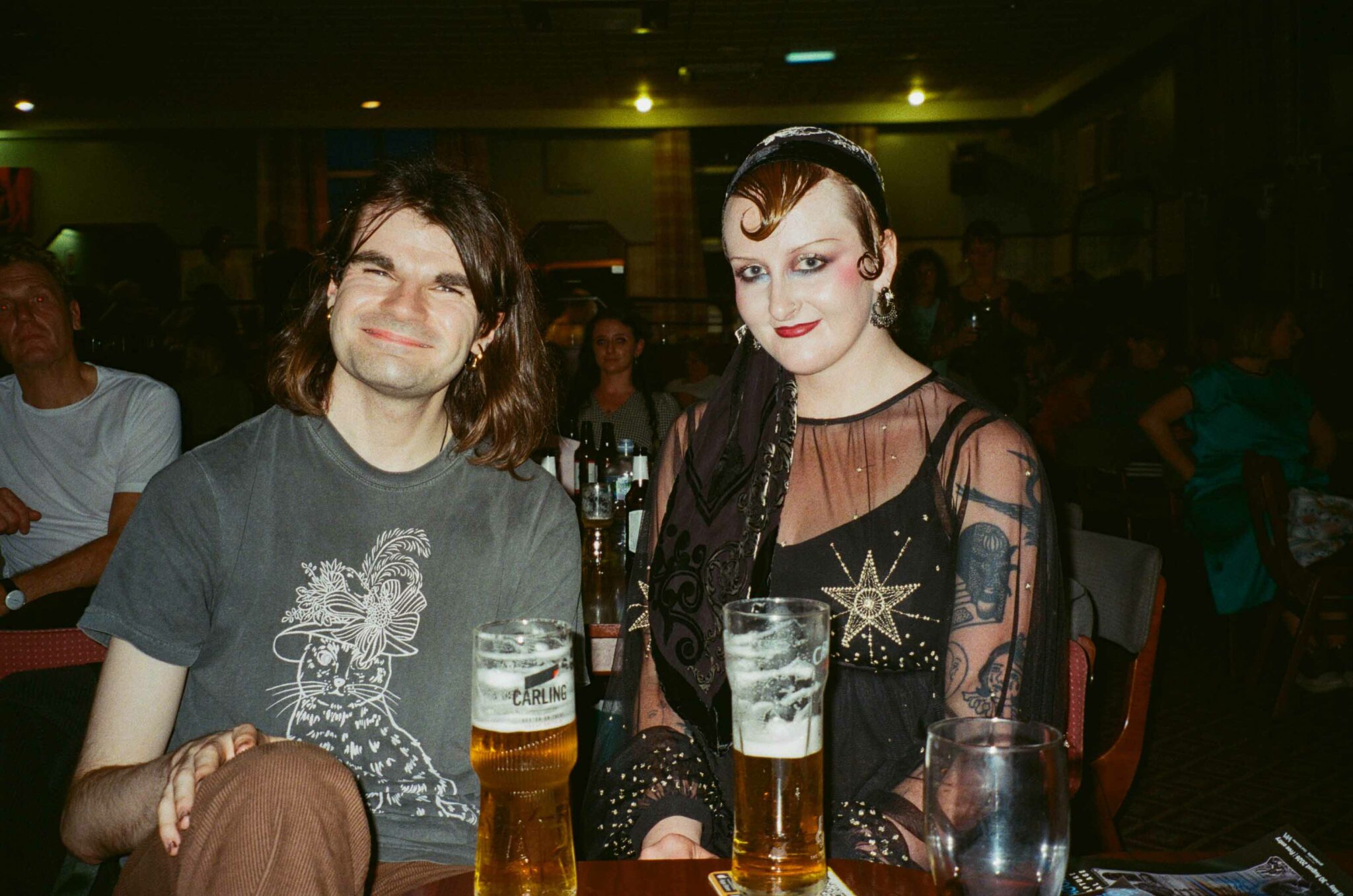 Two audience members sit together at a table, smiling towards the camera with drinks in front of them. One wears a black outfit with sheer sleeves and bold makeup, capturing the expressive style and welcoming atmosphere of the community cabaret event.