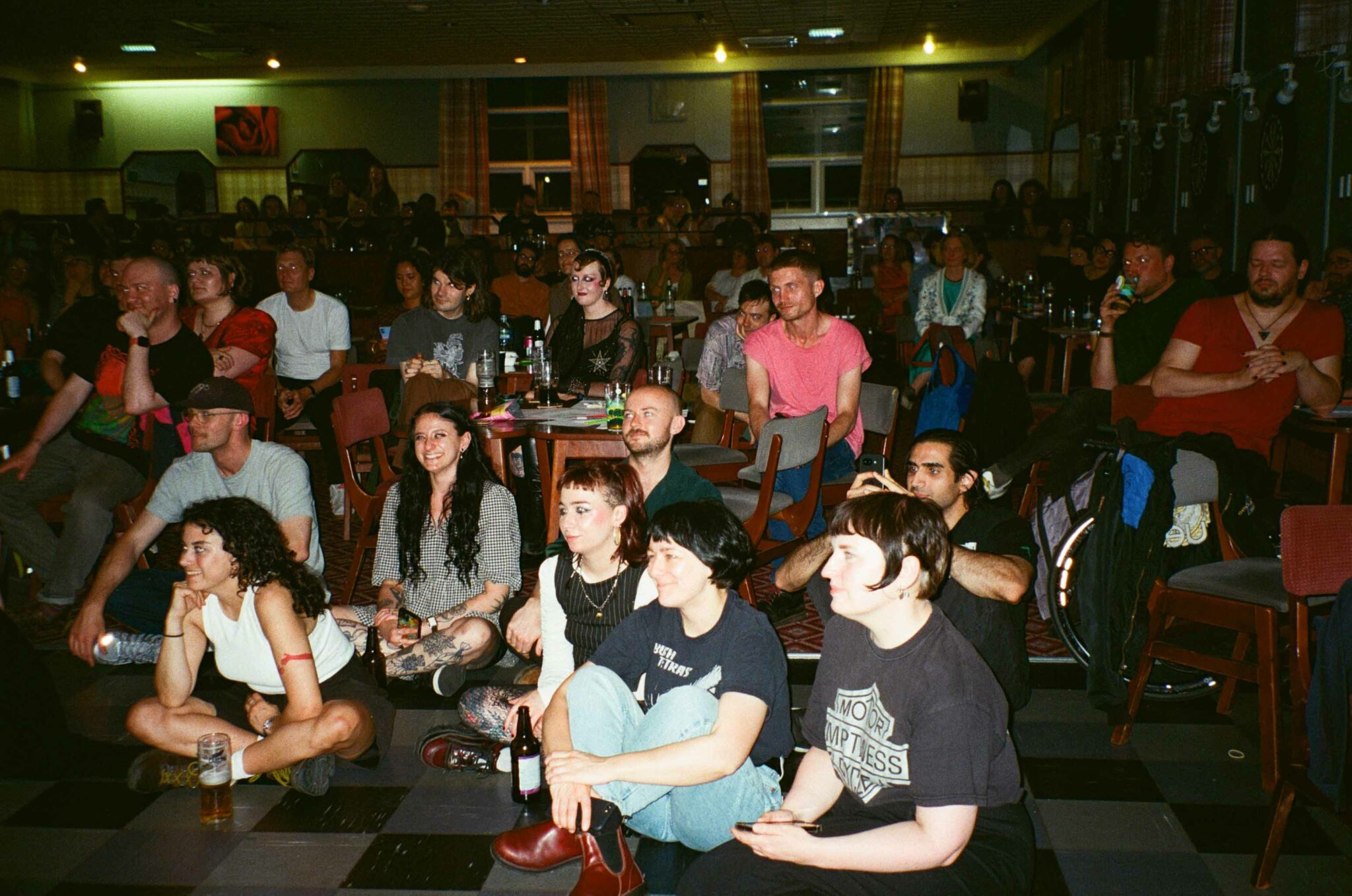 Audience members sit at tables and on the floor in a community hall, watching a live performance with smiles and attentive expressions. Drinks and small tables are scattered throughout the room, creating an informal, welcoming atmosphere for a community cabaret or arts event.