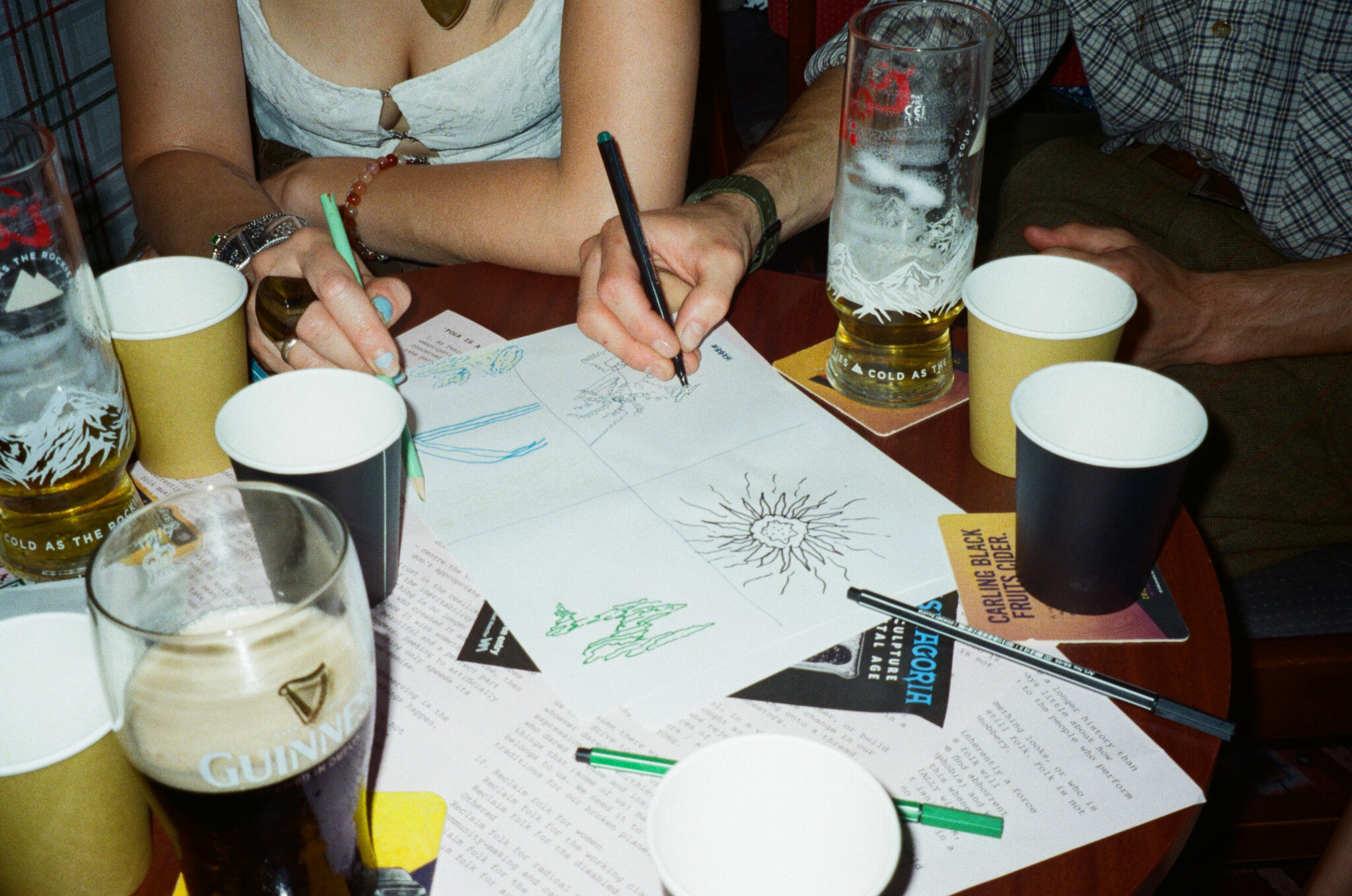 Close-up of a table where several people are drawing together on a large sheet of paper with coloured pens. Surrounded by paper cups, glasses, drinks mats and printed pages, the scene captures a relaxed, collaborative creative activity in a pub or community venue.