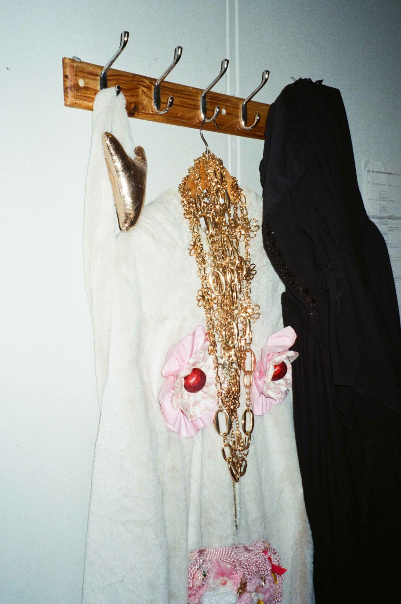 A white faux-fur costume hangs from wooden coat hooks in a backstage dressing room, adorned with layered gold chains, pink ruffles and metallic accessories. A black garment hangs beside it, highlighting the elaborate costumes and preparations behind the performance.