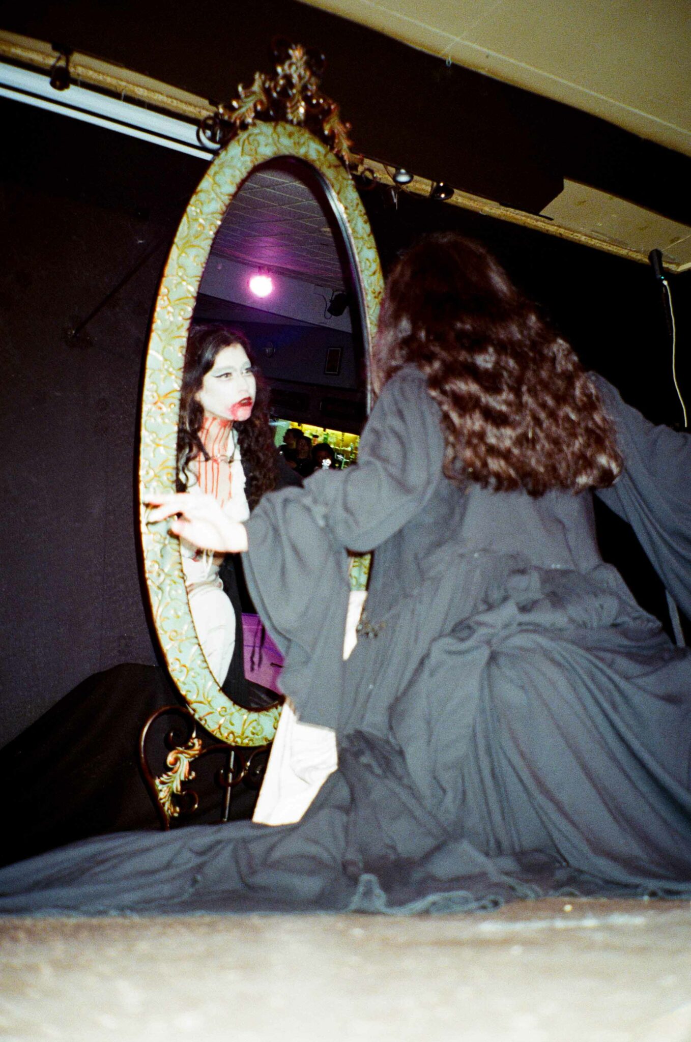 performer in flowing black costume stands before an ornate freestanding mirror on stage, with their reflection revealing dramatic white face makeup and vivid red details around the mouth and neck. The image captures a theatrical moment of transformation and self-reflection during a live performance.