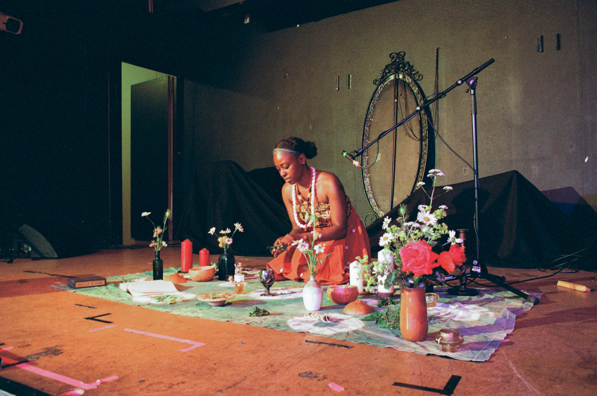 A performer kneels on a stage beside an arrangement of flowers, candles, bowls and other ceremonial objects. A microphone stand and decorative mirror frame the intimate performance setting.