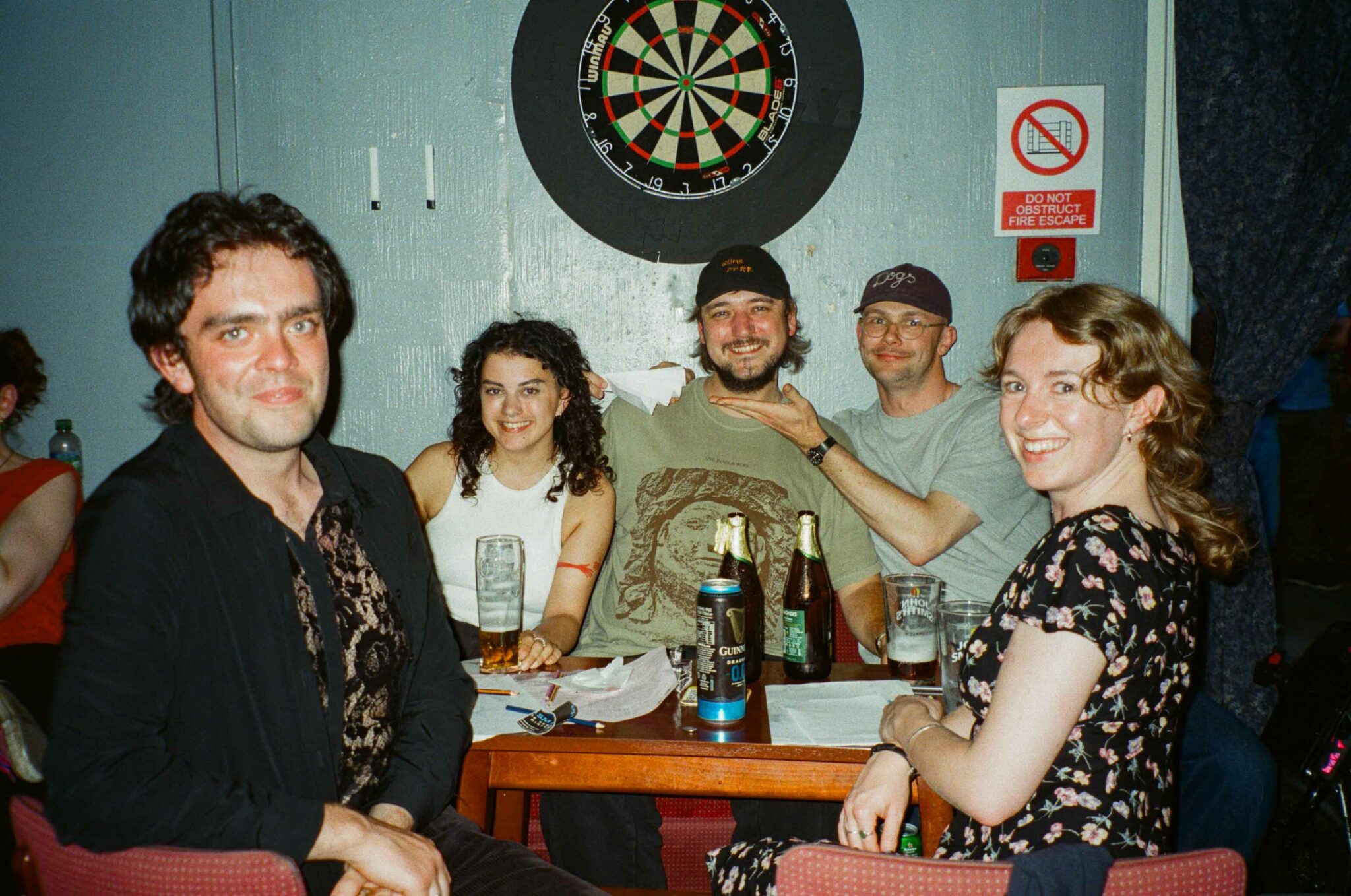 Five audience members sit around a table in a community hall, smiling towards the camera during a cabaret event. Drinks, notebooks and paper cups are spread across the table, while a dartboard on the wall behind them reinforces the relaxed, social atmosphere of the community venue.