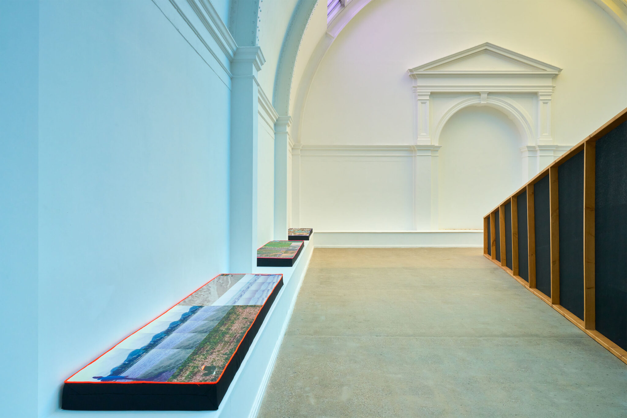 Wide view of a gallery installation with a series of textile-covered benches along one wall and a large wooden-framed structure opposite, beneath high arched architecture.