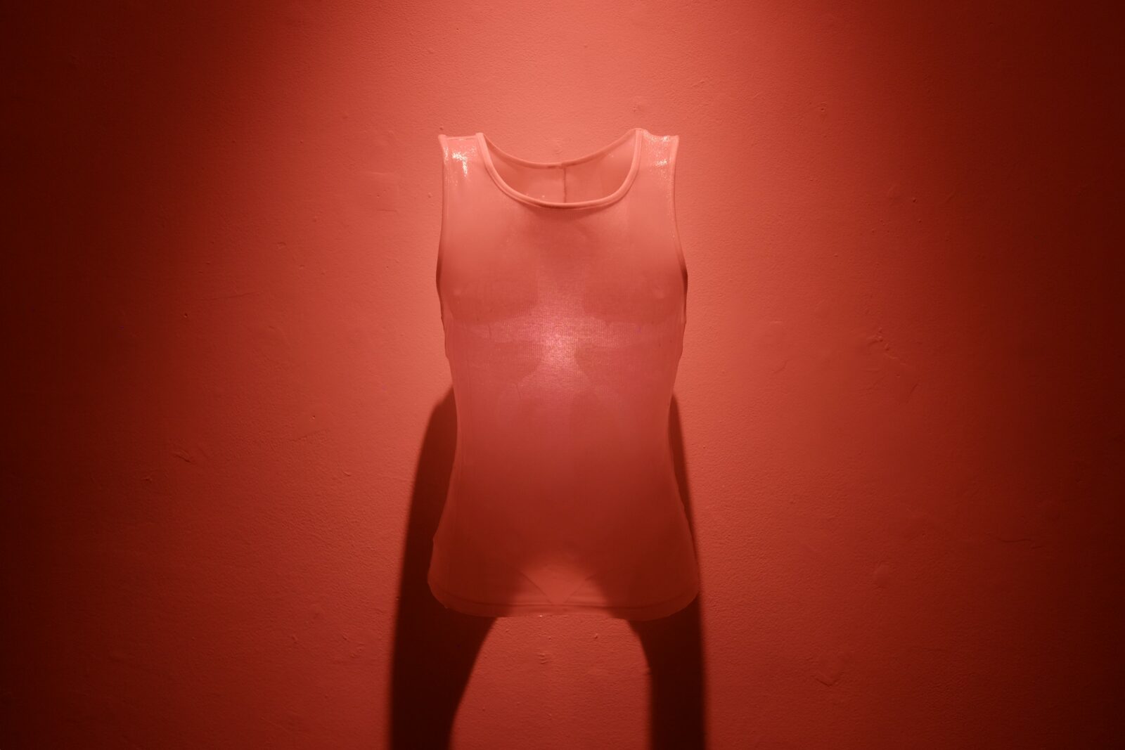 A translucent sleeveless vest hangs against a deep red wall illuminated by red light.