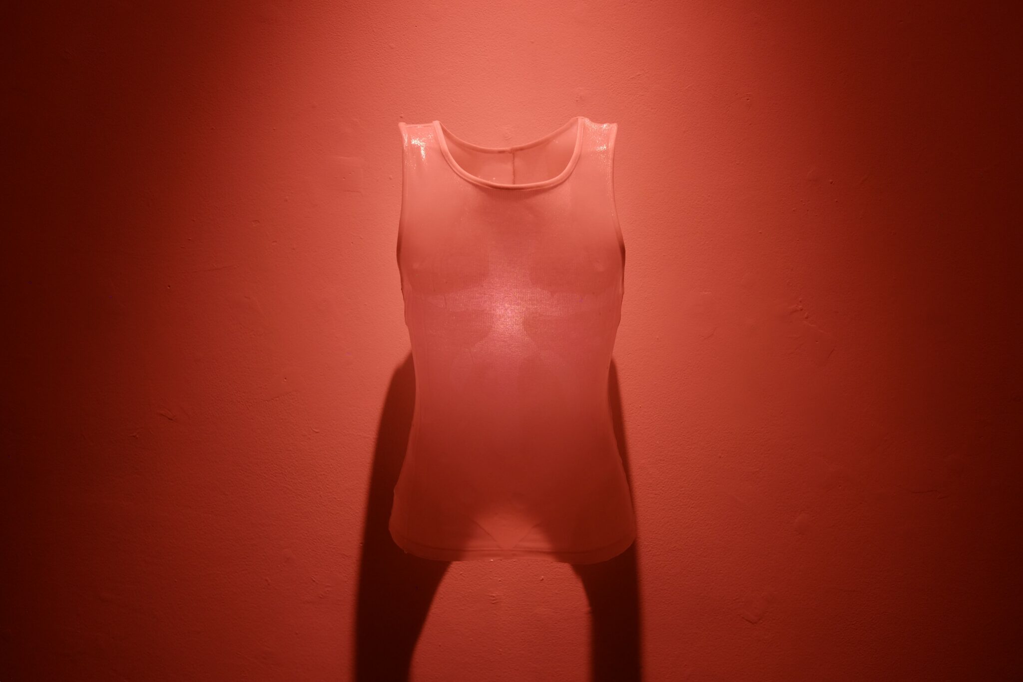 A translucent sleeveless vest hangs against a deep red wall illuminated by red light.